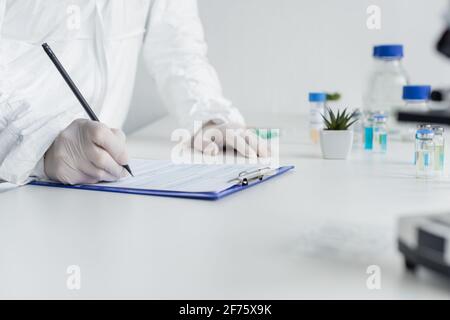 Beschnittene Ansicht des Wissenschaftlers in einem Hazmat-Anzug und Latexhandschuhen Schreiben auf Zwischenablage in der Nähe von Impfstoffen auf verschwommenem Vordergrund Stockfoto