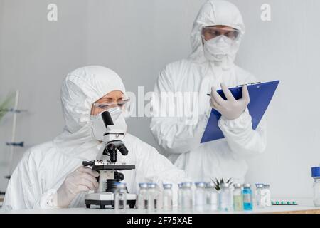 Wissenschaftler in Schutzuniform unter Verwendung des Mikroskops in der Nähe von Kollegen und Impfstoffen Im unscharfen Vordergrund Stockfoto