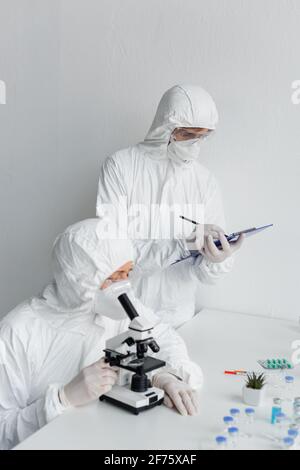 Wissenschaftler in Hazmat Anzug Erforschung Impfstoff in der Nähe von Kollegen schriftlich auf Zwischenablage im Labor Stockfoto