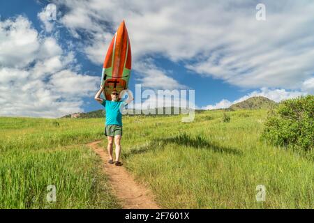 Ältere Paddler tragen ein langes Stand Up Paddleboard (SUP) bergab auf einem Trail im Frühsommer - Lory State Park, Colorado Stockfoto