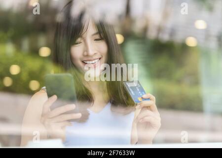 Asiatische Frau mit Kreditkarte mit Handy für Online-Shopping in modernen Coffee Shop, Technologie Geldbörse und Online-Zahlungskonzept, Kredit Stockfoto
