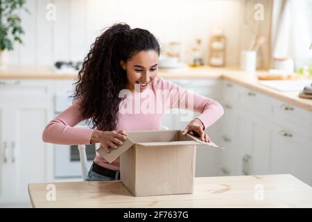 Aufgeregt glücklich junge Frau Auspacken Papppaket in der Küche Stockfoto