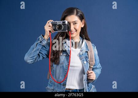 Glückliche junge asiatische Frau halten und schießen Foto mit Vintage-Kamera auf blauem Hintergrund, tragen Backpacker und Gepäck, Urlaub und Reise Konz Stockfoto