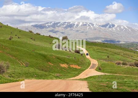 Blick auf den Mount Hermon mit einem schneebedeckten Gipfel in den Wolken. Straße mit dem Auto zwischen den grünen Hügeln Stockfoto