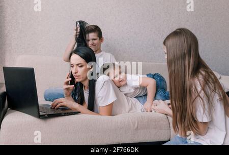 Aktive Kinder frönen, stören und ablenken ihre Mutter, während sie zu Hause auf einem Laptop arbeiten. Während einer Pandemie arbeiten. Freiberuflicher Telearbeit Stockfoto