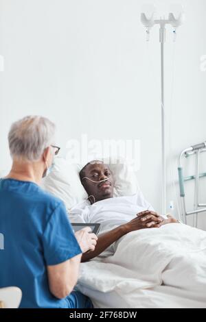 Vertikales Porträt eines jungen afroamerikanischen Mannes, der im Krankenhaus liegt ed Und im Gespräch mit dem Oberarzt Stockfoto