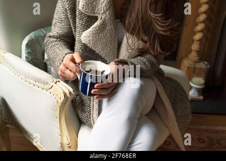 Nahaufnahme einer Frau in warmer Strickmode, die mit einer Tasse Kaffee auf einem Ledersessel sitzt. Indoor Freizeit Lifestyle mit rustikalem Landhausstil. London, Großbritannien Stockfoto