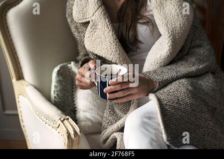 Nahaufnahme einer Frau in kuscheligem warmen Mantel, die auf einem Vintage-Sessel sitzt und eine Tasse Kaffee hält. Indoor-Lifestyle mit entspannter Country-Atmosphäre. London, Großbritannien Stockfoto