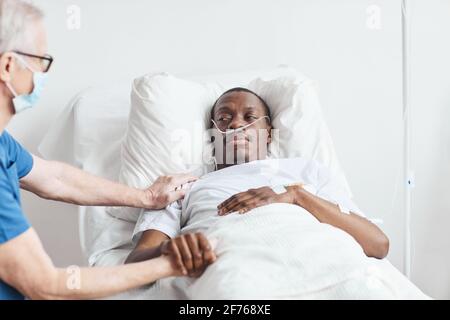 Porträt eines afroamerikanischen Mannes, der im Krankenhausbett liegt, während ihn ein leitender Arzt tröstet, Kopierraum Stockfoto