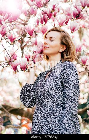 Eine attraktive kaukasische Frau steht an einem sonnigen Frühlingstag bei einem blühenden Magnolienbaum. Stockfoto