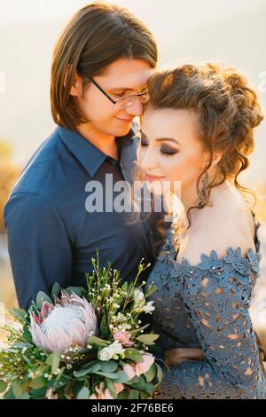 Die Braut und der Bräutigam umarmen sich zärtlich, die Braut hält ein Bouquet mit protea, Eukalyptus und Rosen in den Händen, aus nächster Nähe Stockfoto