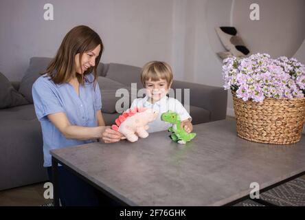 Mama und Kleinkind Sohn spielen Rollenspiele mit gefüllten Spielzeug Dinosaurier auf dem Tisch. Fröhliche Interaktion, Spiele mit Kindern, glückliche Kindheit, Mutter Stockfoto