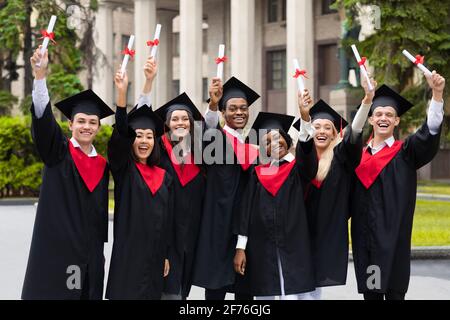 Fröhliche multirassische Studenten in Abschlusskostümen, die Diplome anheben Stockfoto