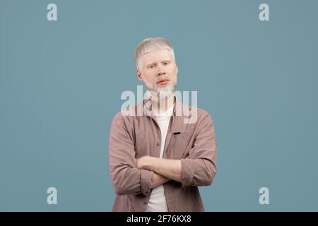 Junger Albino-Mann posiert mit gekreuzten Armen und schaut mit Ernsthafter Gesichtsausdruck bei der Kamera auf türkisfarbenem Hintergrund Stockfoto