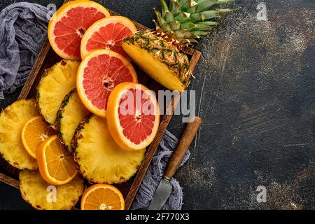 Geschnittene reife Ananas, Orange und Grapefruit in Holzkiste auf dunkelbraunem Steingrund. Tropische Früchte. Draufsicht. Freier Speicherplatz für Text. Modell. Stockfoto