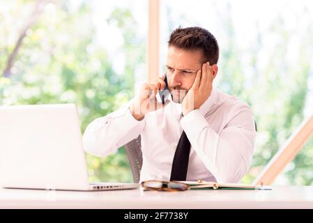 Aufnahme eines besorgten Geschäftsmannes, der mit jemandem auf seinem Mobiltelefon sprach, während er hinter ihrem Laptop am Schreibtisch saß. Stockfoto