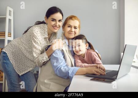 Die kleine Enkelin und ihre Mutter umarmen ihre Großmutter, die zu Hause auf einem Laptop arbeitet. Stockfoto