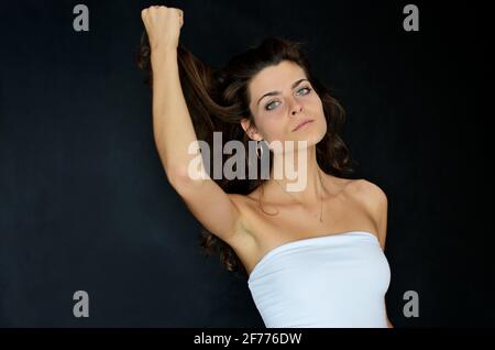 Junge polnische Frau im Studio mit schwarzem Hintergrund. Sie hält ihre Haare. Schlanke Frau mit dunklen Haaren, mit weißem Oberteil und blauer Jeans. Stockfoto