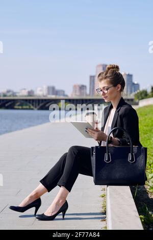 Junge beschäftigt blonde Frau in formalwear trinken und verwenden Tablet beim Sitzen vor dem Flussufer in urbaner Umgebung An einem sonnigen Tag Stockfoto