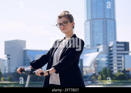 Junge blonde Geschäftsfrau in formalwear und Brillen stehen auf elektrisch Scooter gegen moderne Architektur an sonnigen Tagen Stockfoto