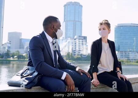 Zwei junge interkulturelle Geschäftspartner in formalwear und Schutzmasken Sitzen in einer städtischen Umgebung, während Sie über die Arbeitspunkte sprechen Stockfoto