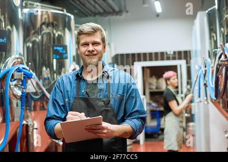 Junger bärtiger Arbeiter einer großen zeitgenössischen Brauerei, der Notizen macht Beim Stehen gegen weibliche Brauerin mit Verarbeitungsmaschine Stockfoto