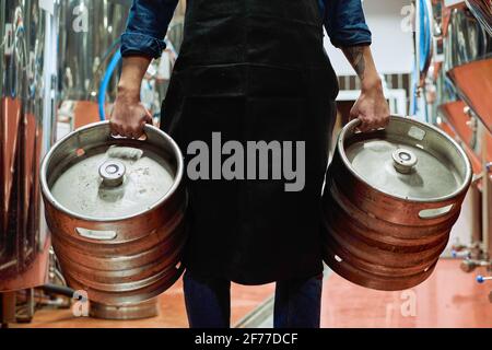 Mittelteil des männlichen Arbeiters der Bierfabrik im Vorfeld Zwei Zisternen tragen, während sie sich vor der Kamera bewegen Reihen von riesigen Stahltanks Stockfoto