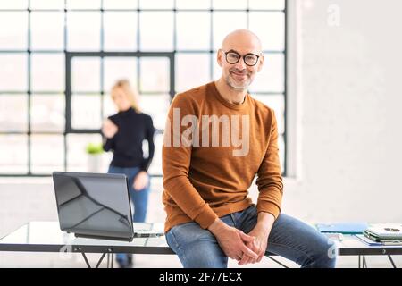 Portait Aufnahme eines lächelnden Geschäftsmannes, der die Kamera ansah und legere Kleidung trug, während er am Schreibtisch im Büro saß. Stockfoto