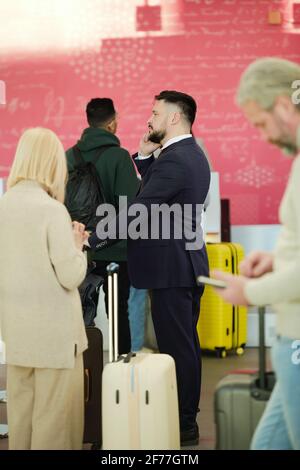 Ein moderner Geschäftsmann in elegantem Anzug, der sich mit dem Mobiltelefon unterhält Vor dem Flug im Flughafen in der Warteschlange für die Registrierung stehen Stockfoto