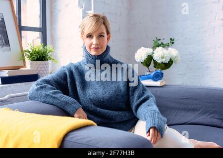 Porträtaufnahme einer schönen blonden Frau, die einen Rollkragenpullover trägt, während sie sich auf der Couch zu Hause entspannt. Stockfoto