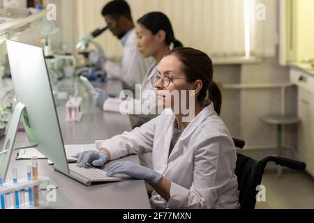 Junge Frau im weißen Mantel sitzt am Tisch Vorderseite des Computermonitors und Arbeiten am Computer im Labor Stockfoto