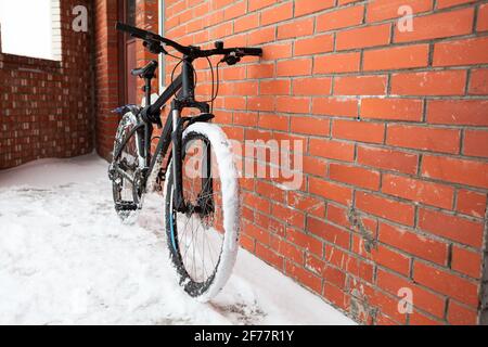 Modernes Mountainbike, das sich an einer roten Backsteinmauer lehnt, schneebedeckte Räder, Winterzeit Stockfoto