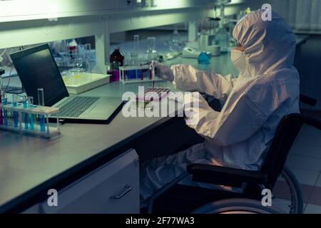 Behinderter Chemiker in Schutzkleidung und Maske, die Flüssigkeit in untersucht Teströhrchen am Tisch im Labor Stockfoto