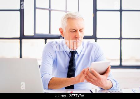 Aufnahme eines Geschäftsmanns, der ein digitales Tablet in der Hand hält, während er am Schreibtisch sitzt und im Büro arbeitet. Stockfoto