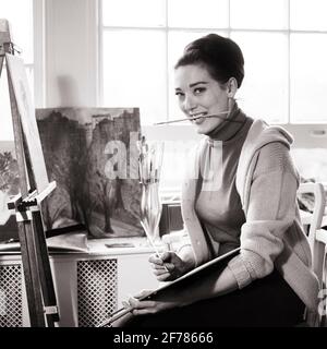 1960ER 1970ER JAHRE KÜNSTLERIN MALEREI AN STAFFELEI LÄCHELND HALTEN FARBE PINSEL IM MUND BLICK AUF DIE KAMERA - A8268 HAR001 HARS ENTSPANNEND FREUDE LEBENSSTIL ZUFRIEDENHEIT FRAUEN ARBEITSPLÄTZE LÄNDLICHEN HAUSLEBEN STAFFELEI KOPIEREN RAUM HALBE LÄNGE DAMEN PERSONEN KÜNSTLER MALER BERUF HANDWERK VERTRAUEN S & W BLICKKONTAKT KÜNSTLER BRÜNETTE VISION GESCHICKLICHKEIT AKTIVITÄT UNTERHALTUNG TRÄUME BERUF GLÜCK FÄHIGKEITEN FRÖHLICH KONTAKT HOBBY INTERESSE KARRIERE HOBBYS WISSEN ZEITVERTREIB ARBEIT STOLZ VERGNÜGEN BESCHÄFTIGUNG BERUFE LÄCHELT PINSEL KÜNSTE FANTASIE FREUDE AN DER ARBEIT DER MITARBEITER KREATIVITÄT MITTLER ERWACHSENER MITTLER ERWACHSENER ENTSPANNUNGSTALENT FÜR FRAUEN Stockfoto