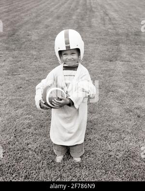 KLEINER JUNGE MIT EINEM LÄCHELN AUS DEN 1960ERN, DER FUSSBALL IN ÜBERGROSSEN HEMDEN HÄLT HELM STEHT IM GRAS UND SCHAUT AUF DIE KAMERA - B11411 LEF001 HARS ERFREUT FREUDE GEWINNEN GESUNDHEIT ZU HAUSE LEBEN KOPIEREN RAUM MENSCHEN KINDER VOLLE LÄNGE KÖRPERLICHE FITNESS MÄNNER SELBSTVERTRAUEN AUSDRÜCKE S & W AUGENKONTAKT GRÖSSE GLÜCK FRÖHLICH HOCHWINKEL MUT AUFREGUNG ÜBERGROSSE ERHOLUNGSMÖGLICHKEIT LÄCHELT KONZEPTUELL FREUDIG STYLISH BABY JUNGE ANGENEHM ANGENEHM CHARMANT WACHSTUM LIEBENSWERT ANGENEHM LIEBENSWERT AMERICAN FOOTBALL ANSPRECHEND SCHWARZ UND WEISS KAUKASISCH ETHNIE ALTMODISCH ZU GROSS Stockfoto