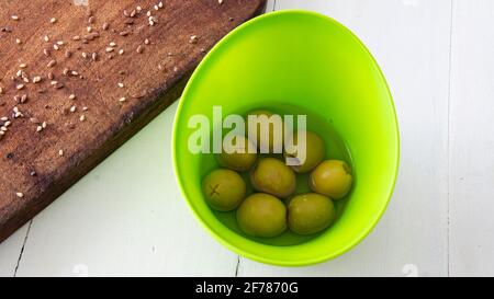 Blick auf eine kleine grüne Schüssel mit grünen Oliven in Salzlake im Inneren. Auf einem Tisch platziert Stockfoto