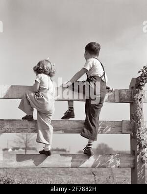 RÜCKANSICHT VON JUNGEN UND MÄDCHEN, BRUDER UND SCHWESTER AUS DEN 1940ER JAHREN BEIDE TRAGEN FARM OVERALLS KLETTERN HÖLZERNE DREI BRETT WEIDEZAUN - J405 HAR001 HARS LIFESTYLE ZUFRIEDENHEIT FRAUEN BRÜDER LÄNDLICHE GESUNDHEIT HOME LEBEN KOPIEREN RAUM FREUNDSCHAFT IN VOLLER LÄNGE INSPIRATION OVERALLS LANDWIRTSCHAFT MÄNNER GESCHWISTER SCHWESTERN B&W ERFOLG GLÜCK ABENTEUER BEIDE ENTDECKUNG WEIDE STRATEGIE UND DIE LANDWIRTE AUS DEM NIEDRIGEN WINKEL WERDEN IN DIE RÜCKANSICHT DER CHANCEN VORGERÜCKT GESCHWISTER KONZEPTIONELL AUS DER RÜCKANSICHT WACHSTUM JUGENDLICHE LÖSUNGEN ZWEISAMKEIT SCHWARZ-WEISS KAUKASISCHE ETHNIZITÄT HAR001 ALTMODISCH Stockfoto