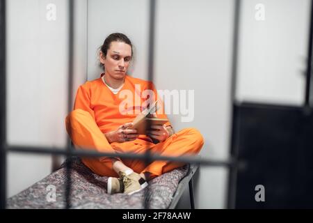 Ein junger Insasse mit tätowiertem Gesicht wird in einer Zelle ferngesteuert trainiert. Liest ein Lehrbuch, das auf einem Koje sitzt Stockfoto