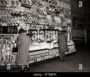 1940ER-JAHRE ANONYME FRAU UND MANN BEI DER AUSWAHL DES EINKAUFS PUBLIKATIONEN BÜCHER UND ZEITUNGEN AN GUT SORTIERTEN ZEITUNGSKIOSKS - R1854 HAR001 HARS DAMEN PERSONEN MÄNNER ZEITUNGEN B&W SHOPPER SHOPPER PENDLER DISCOVERY KUNDENDIENST UND CHOICE AUSSENNEWSSTAND – GELEGENHEIT ZUR RÜCKANSICHT ZEITSCHRIFTEN BERUFE BAHNHOF BEQUEM VON HINTER KIOSK RÜCKANSICHT PAPERBACK SCHWARZ-WEISS CONVENIENCE HAR001 ALTMODISCHE ZEITSCHRIFTEN GEDRUCKT Stockfoto