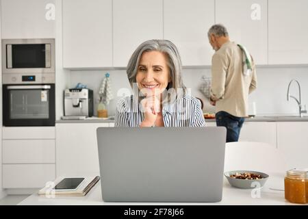 Glückliche reife Frau, die online im Heimbüro arbeitet, Ehemann, der in der Küche kocht. Stockfoto