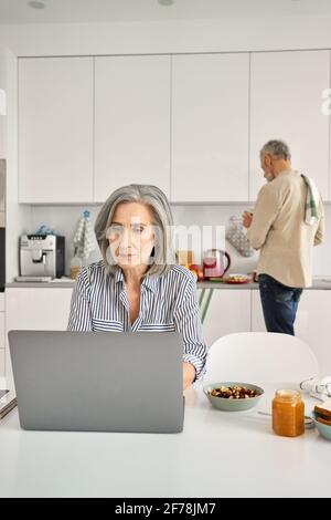 Reife Frau arbeitet online im Home Office, während alter Mann in der Küche kocht. Stockfoto