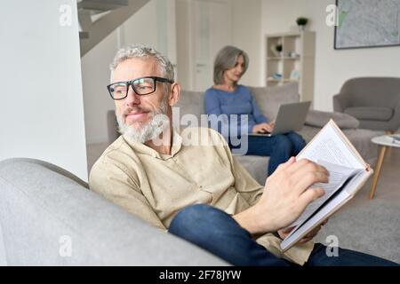 Ein glücklicher älterer Mann, der das Buch liest, entspannt auf der Couch zu Hause sitzen. Stockfoto
