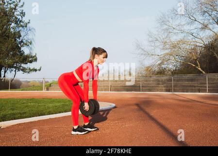 Junge Fitness-Frau in roter Sportkleidung, die im Stadion Trainingseinheiten im Freien macht. Sportlerin, die mit Kurzhanteln arbeitet. Aktiver Lebensstil, Sport Stockfoto