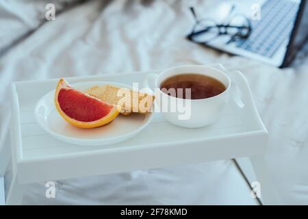 Weißes Holztablett mit einer Tasse heißen Tee oder Kaffee, Grapefruit und Keksen mit einem Laptop auf dem Hintergrund. Frühstück von Geschäftsreisenden. Morgen Stockfoto