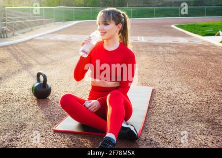 Lächelnde Fitness-Frau trinkt Wasser aus einer wiederverwendbaren Glasflasche und ruht sich während des Trainings im Freien auf einer Matte im Stadion aus. Aktiv Stockfoto