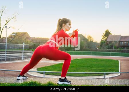 Rückansicht Junge Fitness-Frau im roten Sport trägt beim seitlichen Ausfallschritt, Kniebeugen beim Training im Freien auf der Stadiontreppe. Dehnung nach dem Laufen Stockfoto