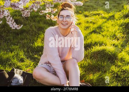 Junge kaukasische Hipster Mädchen in Brille und leichte Kleidung auf grünem Gras in der Nähe blühenden Baum im Park sitzen und mit ihrem Smartphone. Lachende Frau Stockfoto