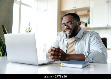 Ein gutaussehender, selbstbewusster, freundlicher, junger afroamerikanischer bärtiger Mann mit Brille und formellem Hemd, ein Geschäftsleiter, Manager oder Broker, der zuhause aus der Ferne arbeitet, schaut auf den Laptop-Bildschirm Stockfoto