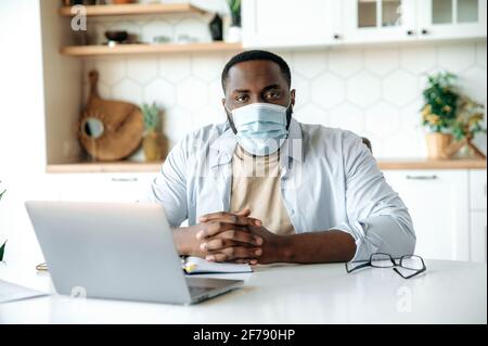 Das Porträt eines jungen afroamerikanischen Mannes in einer medizinischen Maske, in stilvoller Kleidung, der aus der Ferne arbeitet, zu Hause an einem Tisch sitzt, die Kamera anschaut und einen Laptop verwendet. Fernarbeiten während der Quarantäne Stockfoto